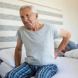 Older man sitting on bed holding his lower back, showing signs of morning back pain common among seniors